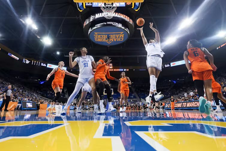 Kiki Rice making a shot in the air as other Oklahoma State and UCLA basketball players surround her, 'MARCH MADNESS' scrolling across the jumbotron