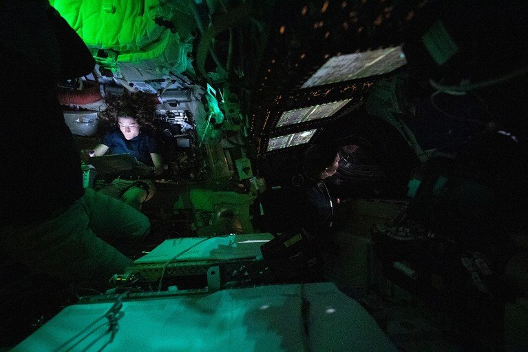 NASA astronaut Christina Koch reads on a tablet in the dimly lit Orion crew capsule as astronaut Jeremy Hansen is seen peering out a window while in space, April 3, 2026.
