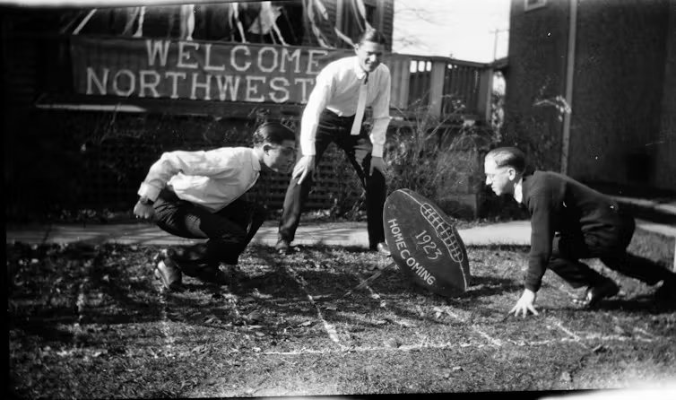A black-and-white photo shows two young men crouching on either side of an oversized football as another young man stands between them.