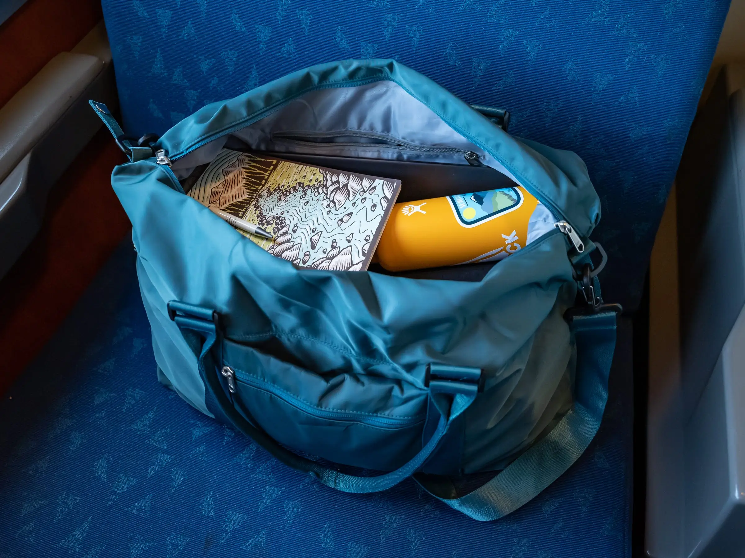 Blue travel bag filled with snacks and orange water bottle on a blue Amtrak seat