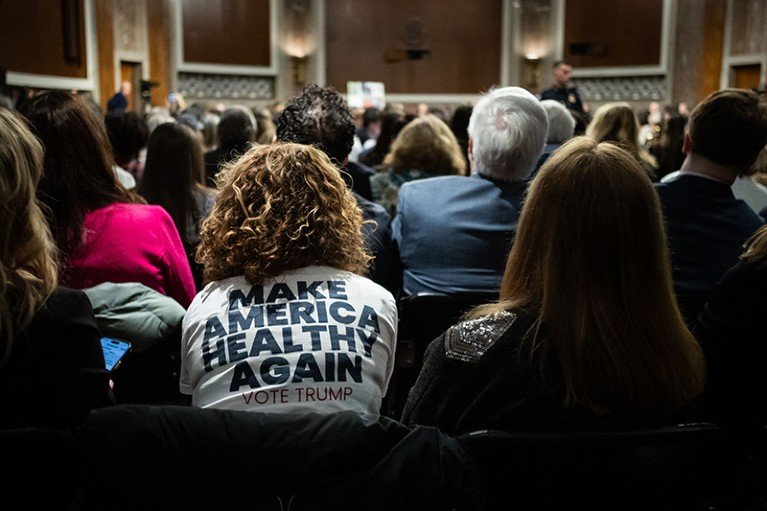 An audience sits in a large senate hearing room, with one attendee wearing a white T‑shirt printed with the slogan “MAKE AMERICA HEALTHY AGAIN — VOTE TRUMP” while a speaker addresses the crowd at the front.