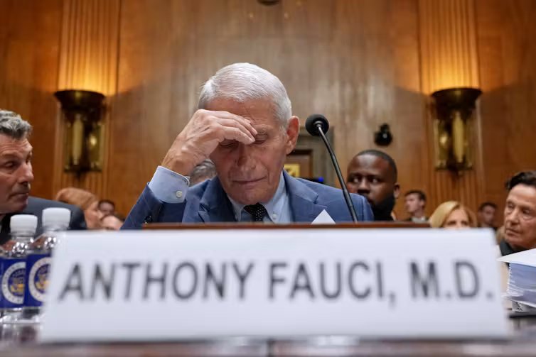 A man in a blue jacket sits behind a nameplate reading Anthony Fauci, M.D., resting his hand against his forehead, a microphone in front of him