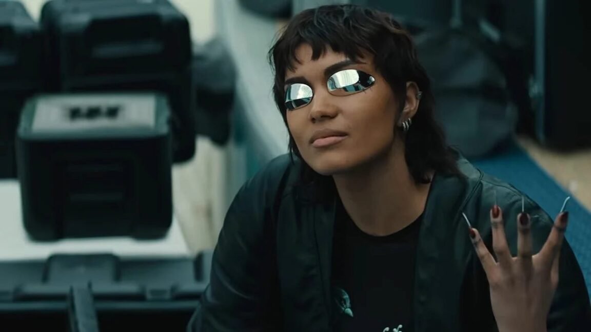 girl with short cropped dark hair wearing futuristic glasses and holding up a hand with long metallic fingernails